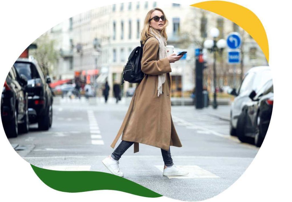 Kobieta w okularach przeciwsłonecznych i długim płaszczu przechodzi przez ulicę, w ręku ma telefon