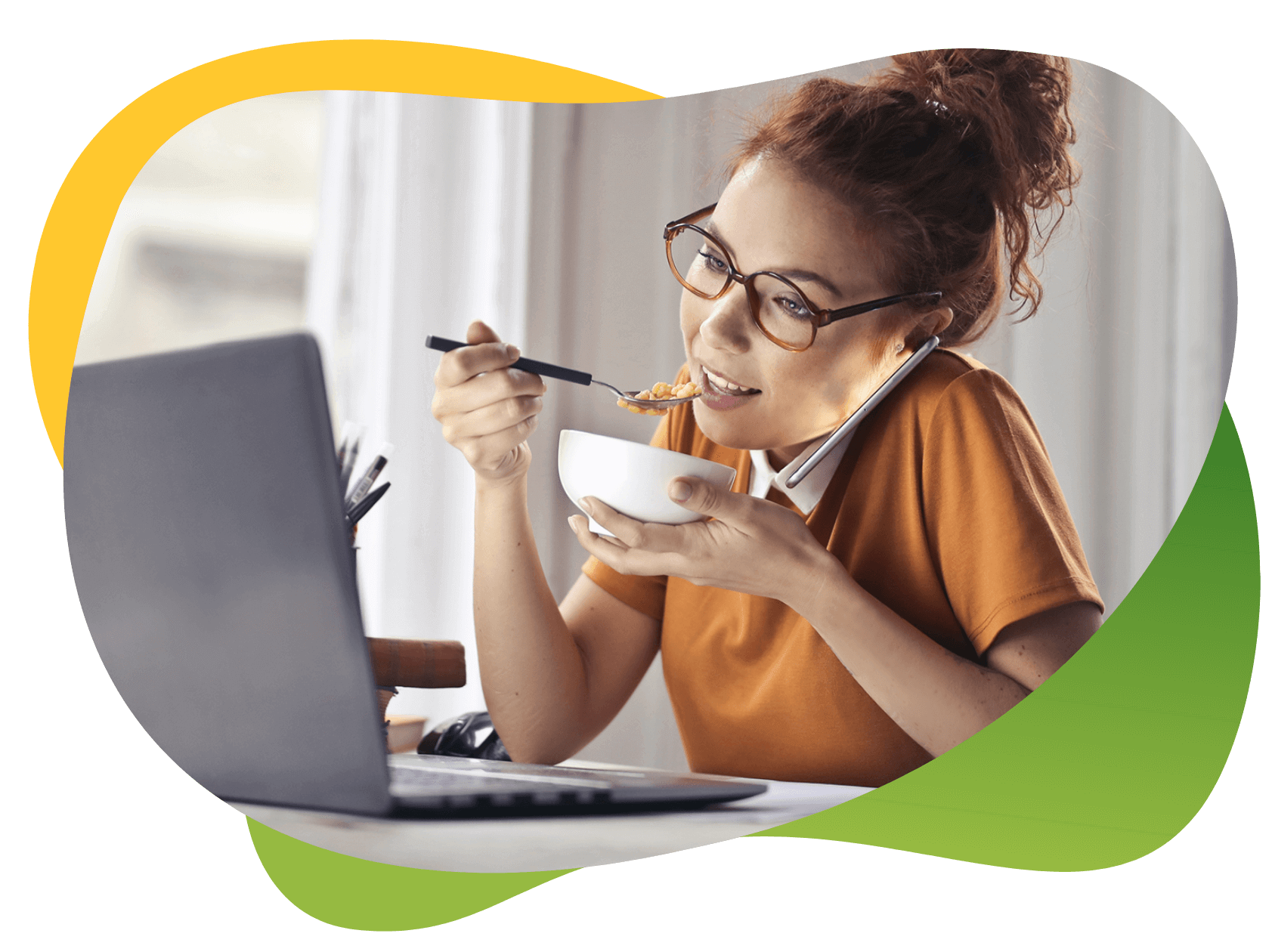 Młoda kobieta w okularach siedzi przed laptopem, jednocześnie rozmawia przez telefon i je posiłek