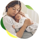 Młoda matka siedzi na sofie z zamkniętymi oczami, przyciska śpiące dziecko do piersi